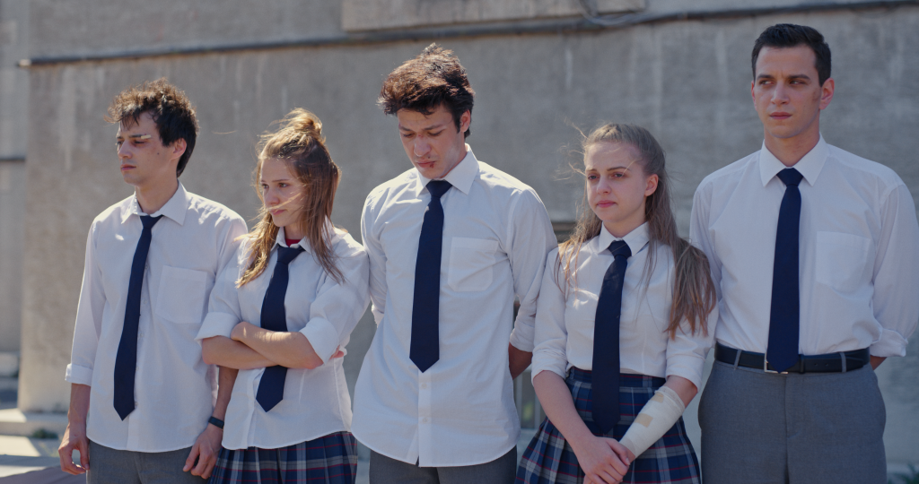 Protagonistas de Love 101 em cena da série; da esquerda para direita: Sinan, Eda, Kerem, Isik e Osman alinhados usando uniformes do colégio com alguns machucados e expressões sérias, decepcionadas e tristes