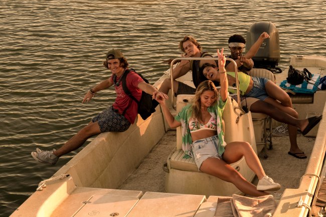 John B., Sarah, Kiara, JJ e Pope em cena de Outer Banks, os cinco estão sorrindo com braços abertos posando dentro de um barco com o mar ao fundo