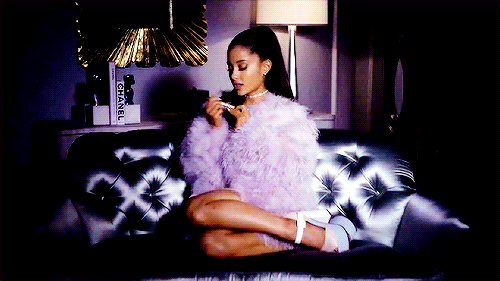 Ariana Grande sentada em um sofá usando um casaco de pelúcia roxo, salto, colar e cabelo preso em um rabo de cavalo lixando a unha.
