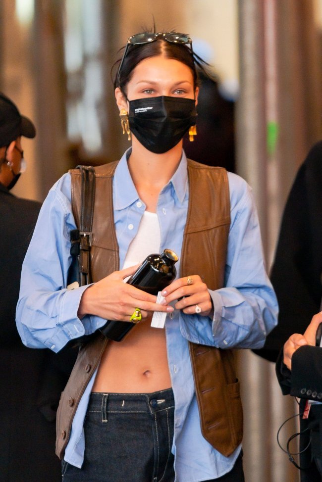 Foto da modelo Bella Hadid na rua. Ela usa um top cropped branco, camisa azul aberta, colete de couro marrom, calça jeans escura, máscara facial preta, óculos de sol no topo da cabeça e cabelo preso em um coque. Ela olha para a frente e não sorri para a foto.