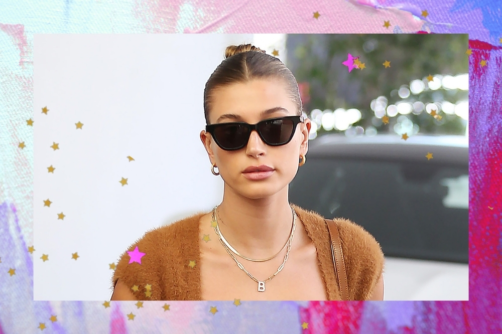 Montagem com a foto da modelo Hailey Bieber com o fundo rosa e roxo e detalhes de estrelinhas douradas. Ela usa um top marrom, óculos de sol, brinco e colares.