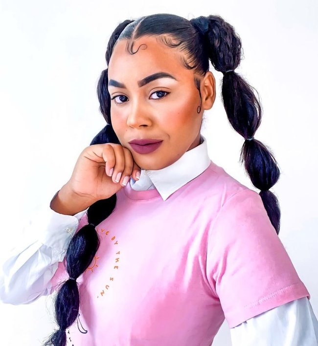 cabelo bubble braid Jovem posando com camisa rosa e branca, olhando par a câmera sorridente com uma das mãos apoiadas no rosto.
