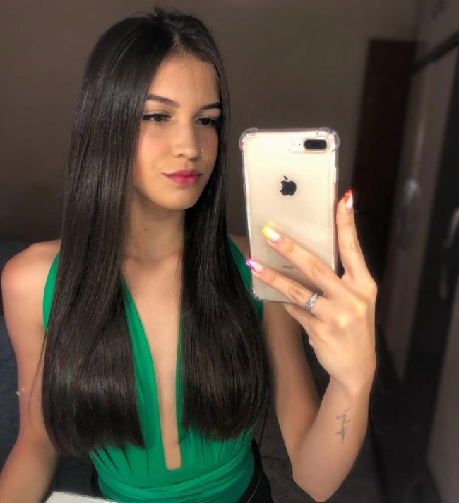 Jovem tirando selfie com blusa verde com decote, segurando celular e com expressão séria.