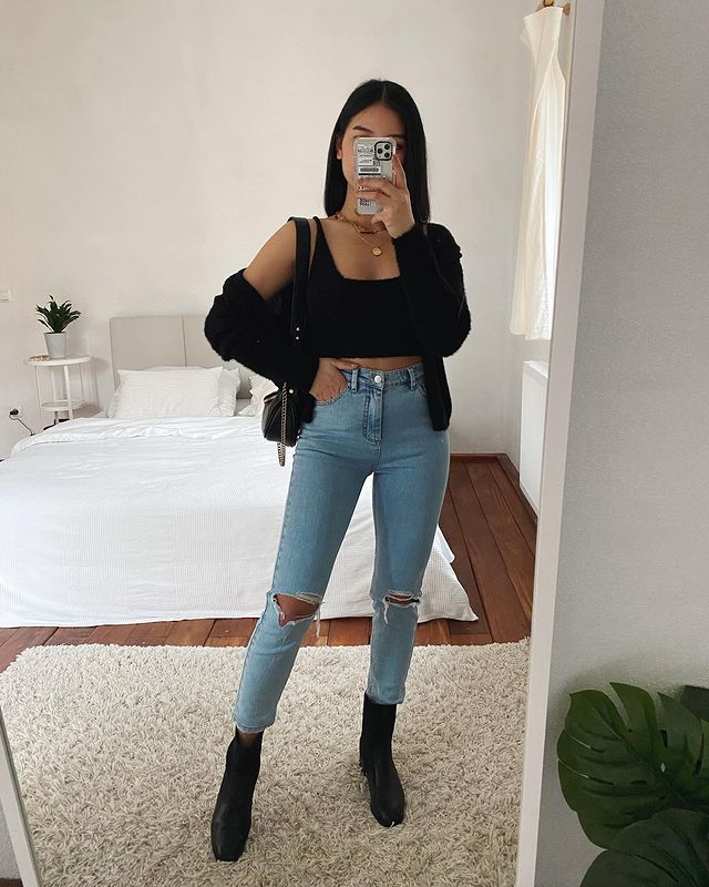 Selfie no espelho de uma mulher. Ela usa um top cropped preto, cardigan preto, calça jeans skinny rasgada nos joelhos, bota preta e bolsa preta.