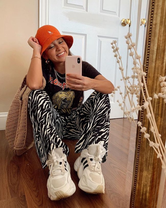 Jovem tirando foto em frente a espelho, ela está sentada e usa bucket hat laranja, camiseta de banda e calça de zebra com tênis branco.