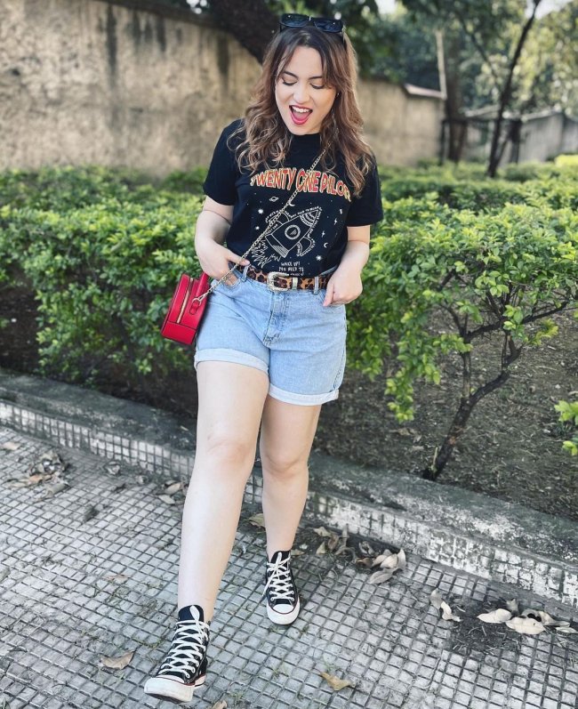 camisa de banda com bolsa e short Jovem posando com as mãos no bolso usando short jeans, all star, bolsa vermelha e camiseta de banda.