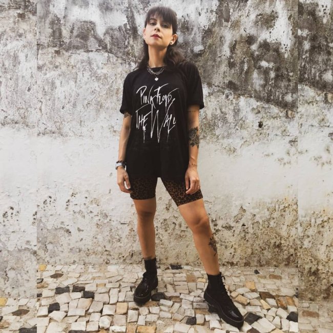 Jovem posando em frente a parede branca com manchas pretas, ela tem uma expressão séria, e usa camiseta de banda com short biker, ambos na cor preta.
