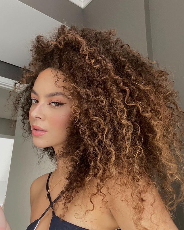 corte cabelo em camadas Selfie de uma mulher. Ela usa um top preto, cabelo crespo solto em um corte em camadas e maquiagem leve. Ela está com o rosto de perfil, olha para a câmera e não sorri.