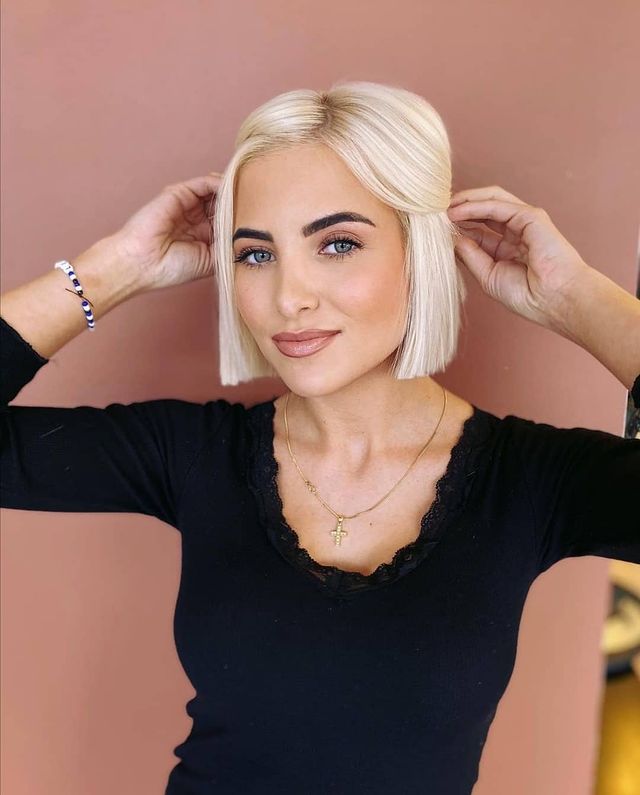 corte chanel cabelo loiro Jovem posando sorridente com as duas mãos apoiadas na cabeça em fundo rosa.
