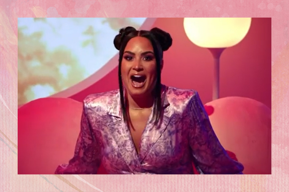 Demi Lovato fazendo expressão de surpresa com a boca aberta e os olhos arregalados; ela está usando um terno em tons de rosa perolado e dois coques com franja lisa e solta; a margem da imagem é uma textura em tons de rosa com branco