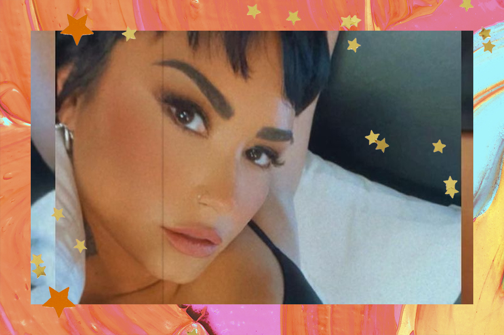 Selfie de Demi Lovato; ela está olhado para câmera com expressão séria e usando um sutiã preto; a margem é uma textura de tintas em tons de laranja, amarelo, rosa, verde e estrelas amarelas e laranjas decoram a imagem