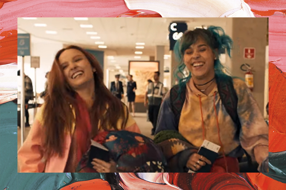 Cena de Diários de Intercâmbio com Larissa Manoela e Thati Lopes; elas estão andando no aeroporto segurando casacos, malas, passagens e passaporte enquanto sorriem; a margem da imagem é uma textura de tintas em tons de rosa, verde, vermelho, laranja e preto