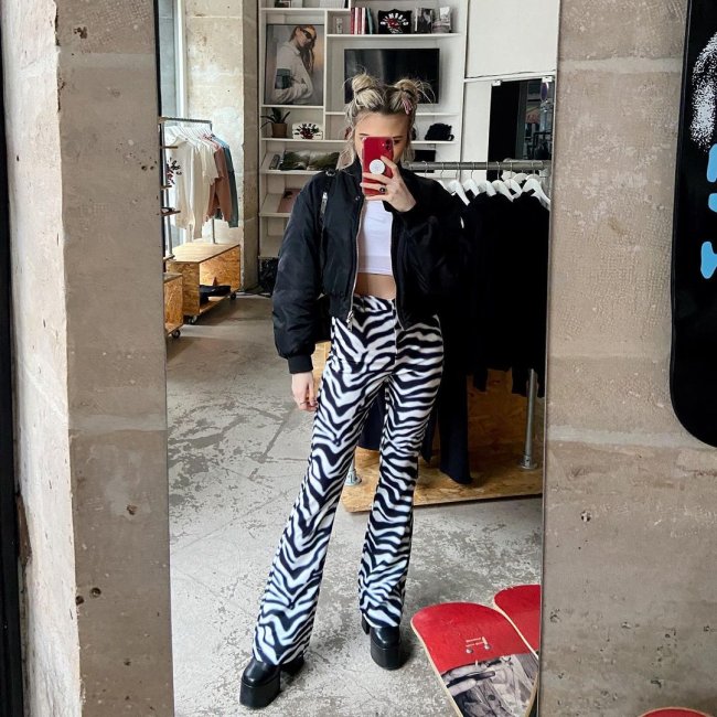 Jovem tirando foto do seu look em frente a espelho. Ela usa jaqueta preta, top branco e calça de zebra.