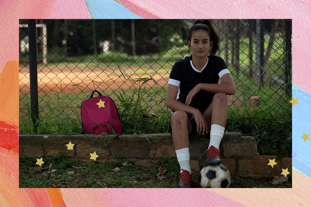 A jogadora de futebol Kezia Sousa está sentada ao lado de um campo, vestido uniforme e com uma bola embaixo do pé