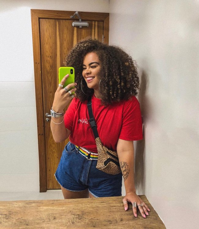 Selfie de uma mulher no espelho. Ela usa camiseta vermelha, short jeans, pochete com estampa de oncinha e cabelo crespo solto com o corte long bob. Ela olha para o celular e sorri.