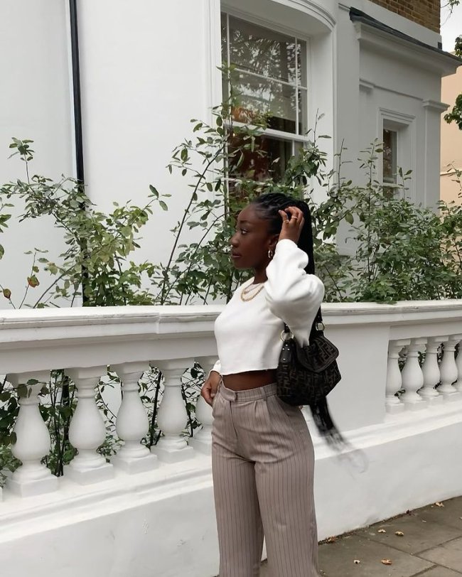 Foto de uma mulher na calçada. Ela usa um moletom branco no modelo cropped, calça de alfaiataria listrada e bolsa preta. Ela está com o cabelo preso em um rabo de cavalo, com a mão esquerda na cabeça, olha para o lado e não sorri.