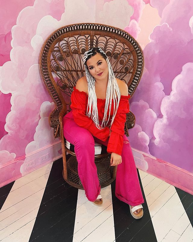 Jovem posando sentada em cadeira de madeira, atrás dela tem uma parede com céu lilás e nuvens brancas, no chão o piso é listrado preto e branco. Ela usa uma camisa vermelha e uma calça rosa.