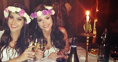 Foto da blogueira Nah Cardoso e cantora Manu Gavassi em um restaurante. Ambas usam uma blusa branca, coroa de flores e seguram uma taça de champanhe sorrindo.