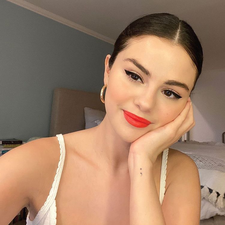 Selfie da atriz e cantora Selena Gomez. Ela usa uma blusa de alcinha branca, brinco de argola dourado, cabelo preso em um coque e maquiagem com delineado preto e batom vermelho. Ela olha para a câmera, sorri e apoia o rosto na mão esquerda.