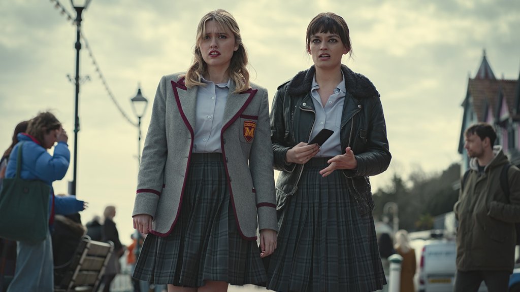 Aimee e Maeve em cena da nova temporada de Sex Education; as duas estão uniformizadas e olhando para frente com expressões confusas; As duas exibem franjinhas como novo visual