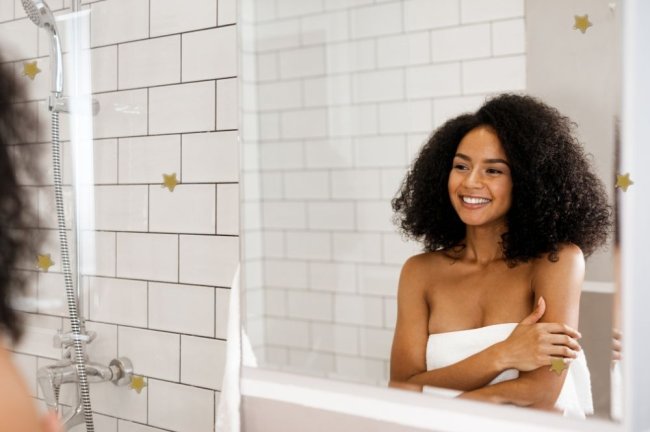 Jovem sorridente enrolada em toalha se olhando no espelho. Ela está dentro de um banheiro branco.