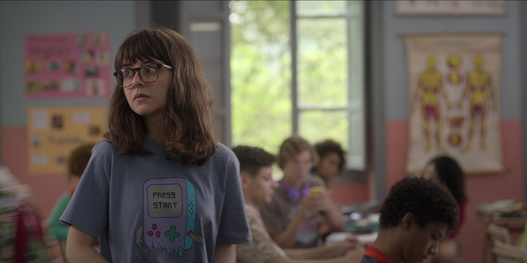Klara Castanho em cena de Confissões de Uma Garota Excluída; ela está usando óculos e camiseta cinza com um videogame de estampa e uma expressão triste