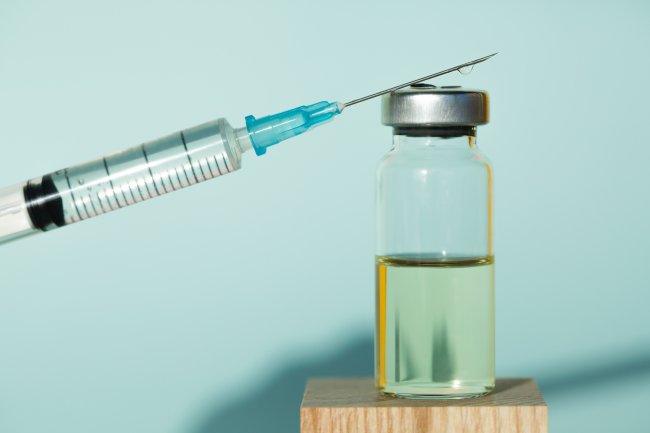 Imagem de um tubo contendo um imunizante sobre um bloco de madeira. Sobre ele, está apoiado uma seringa.