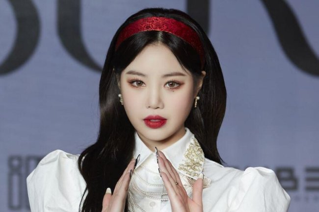 Foto de Soojin usando blusa branca com tiara e batom vermelho; ela está com as mãos levantadas na frente do corpo e tem a expressão séria