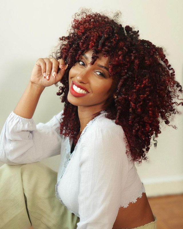 Foto de uma mulher. Ela usa uma blusa de manga bufante branca, maquiagem natural com batom vermelho, cabelo cacheado solto em um tom de vermelho. Ela está sentada em um sofá, olha para a câmera e sorri.
