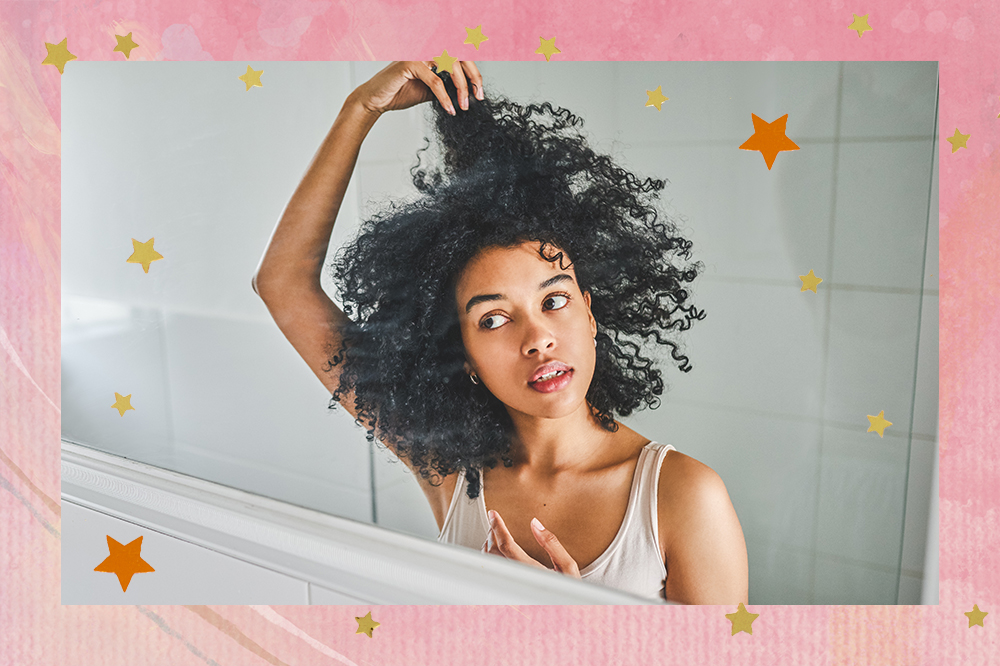 Garota mexendo em seu cabelo crespo em frente a um espelho. A montagem possui fundo rosa e estrelinhas douradas e laranjas em cima.
