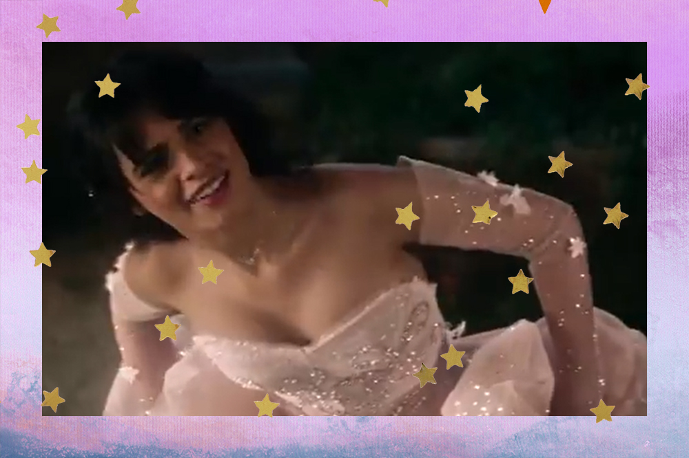 Camila Cabello caracterizada como Cinderela em cena do filme; ela está segurando a barra do vestido se abaixando levemente com uma expressão confusa; a margem é uma textura de degradê roxa com azul e estrelas amarelas decoram a imagem