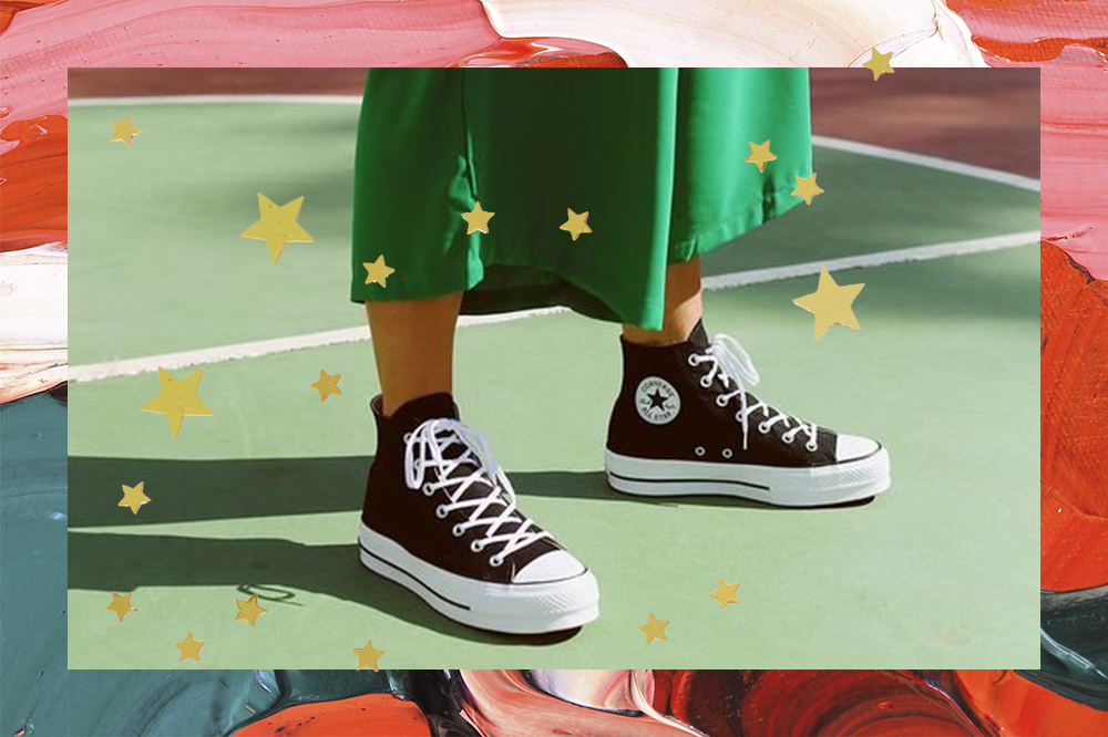 Montagem com foto em fundo de tintas em vermelho, rosa e verde com estrelinhas douradas em cima. Na foto, dá para ver os pés de uma garota usando tênis Chuck Taylor preto e branco da Converse e uma parte da saia midi verde dela.