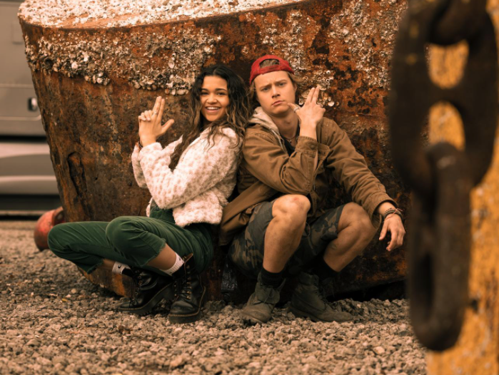 Kiara e JJ juntos sentados encostados em uma pedra com expressões sorridentes.