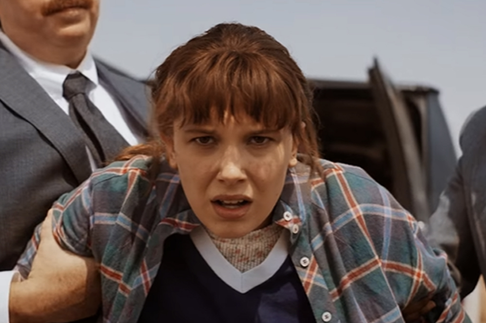 Millie Bobby Brown caracaterizada como Eleven em cena da quarta temporada de Stranger Things; ela está com uma camisa xadrez e uma camiseta preta de gola branca por baixo; a personagem é puxada pelos braços por dois homens e faz uma expressão de inconformada