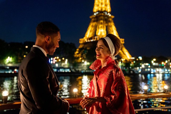 Imagem da segunda temporada de Emily em Paris, série da Netflix; Alfie e Emily estão conversando e sorrindo um para o outro; ele usa um terno preto e ela usa um casaco rosa com o cabelo preso com uma faixa branca; o fundo mostra o rio e as luzes da Torre Eiffel