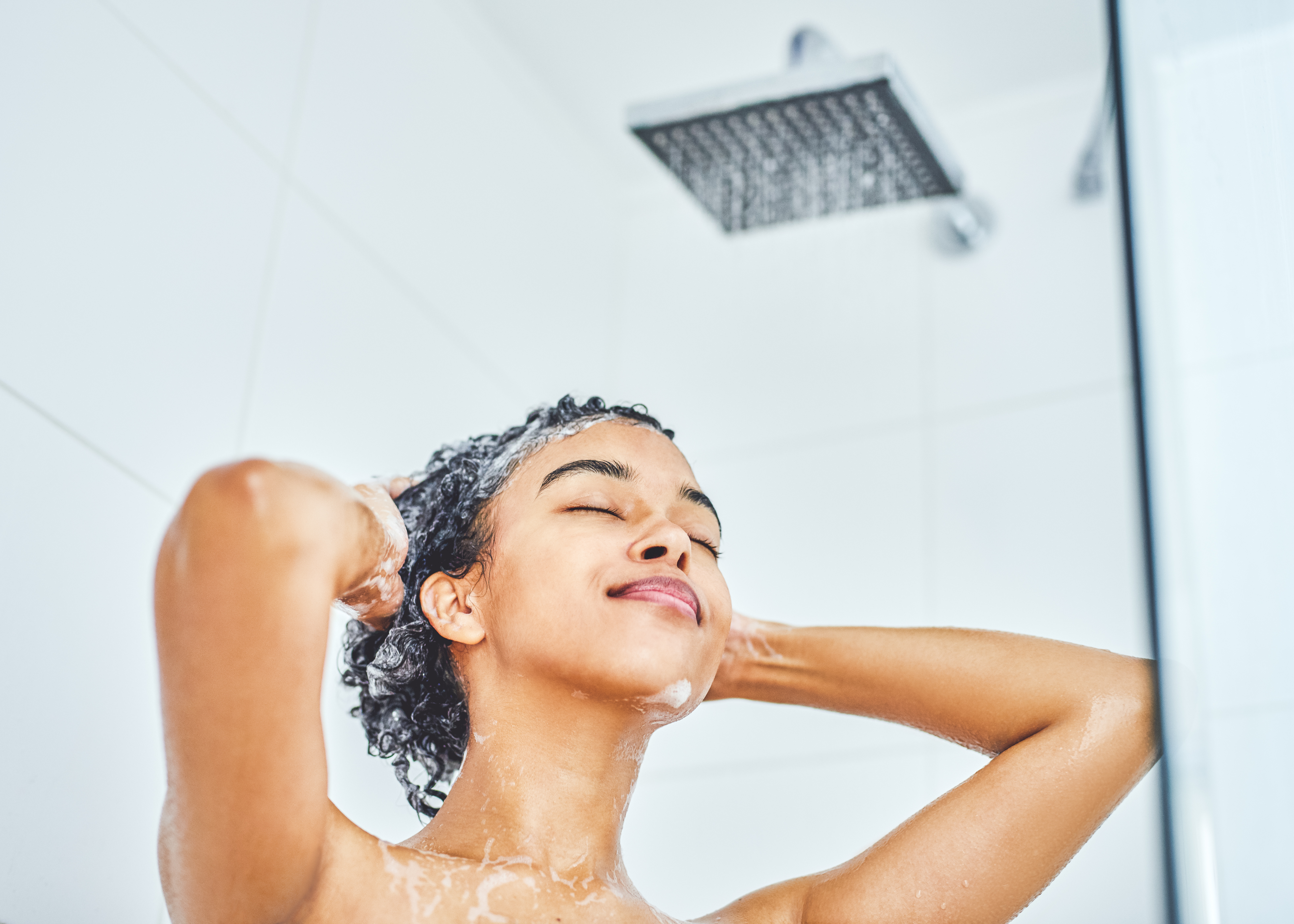 Garota lavando o cabelo no banho. Ela está de olhos fechados, com as duas mãos na cabeça, e na foto de baixo para cima dá para ver o chuveiro.