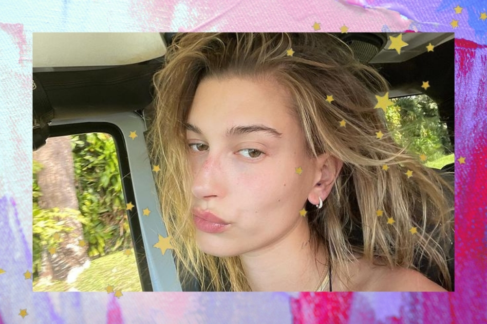 Montagem com uma selfie da Hailey Bieber com o fundo roxo e rosa com detalhes de estrelinhas douradas. Ela está sem maquiagem e com o cabelo molhado e solto dentro de um carro. Ela olha para a foto e faz um bico.