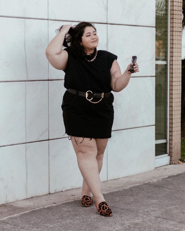 Jovem posando com look preto em frente a parede. Uma de suas mãos está no cabelo.