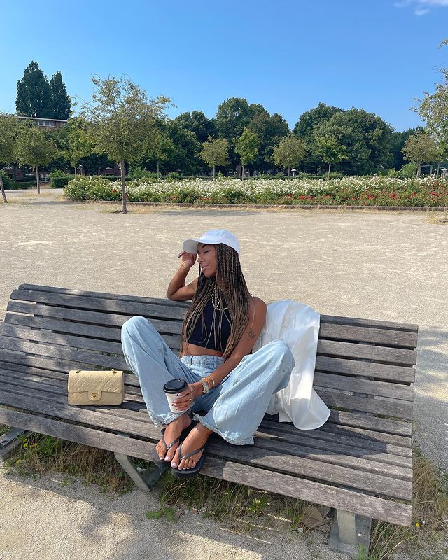 Foto de uma mulher em uma praça sentada em um banco de madeira. Ela usa uma regata cropped preta, calça jeans oversized, chinelo, boné branco e bolsinha. Ela olha para o lado e segura um copo de café na mão esquerda.