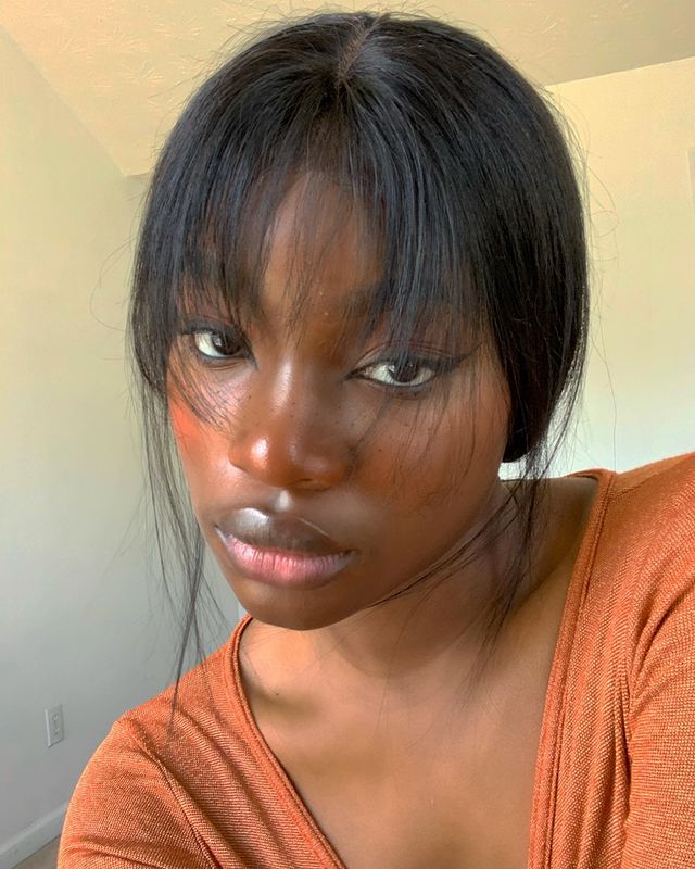Selfie de uma mulher. Ela usa uma blusa laranja, cabelo meio preso com franja repicada solta e maquiagem natural com blush marrom, lápis de olho preto na linha d'água e delineado preto. Ela olha para a câmera e não sorri.