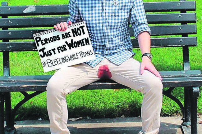 Homem trans sentado no banco de uma praça, com uma calça bege, as pernas abertas e sangue menstrual entre elas