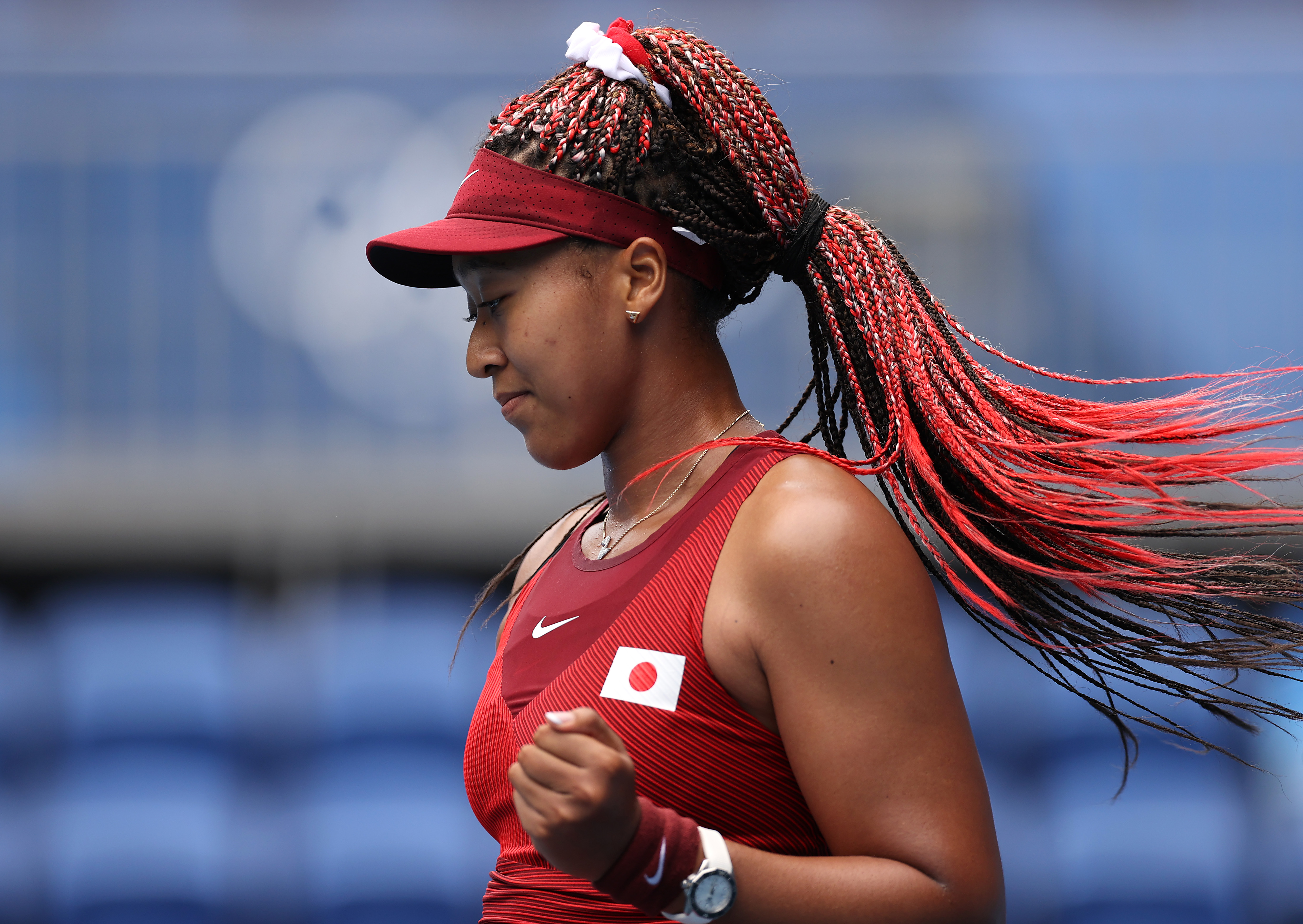 A tenista japonesa Naomi Osaka nas Olimpíadas de Tóquio 2020. Ela está de lado para a câmera, usando viseira vermelha, roupa vermelha, com cabelo trançado em vermelho e comemorando um ponto no jogo com uma das mãos fechadas.