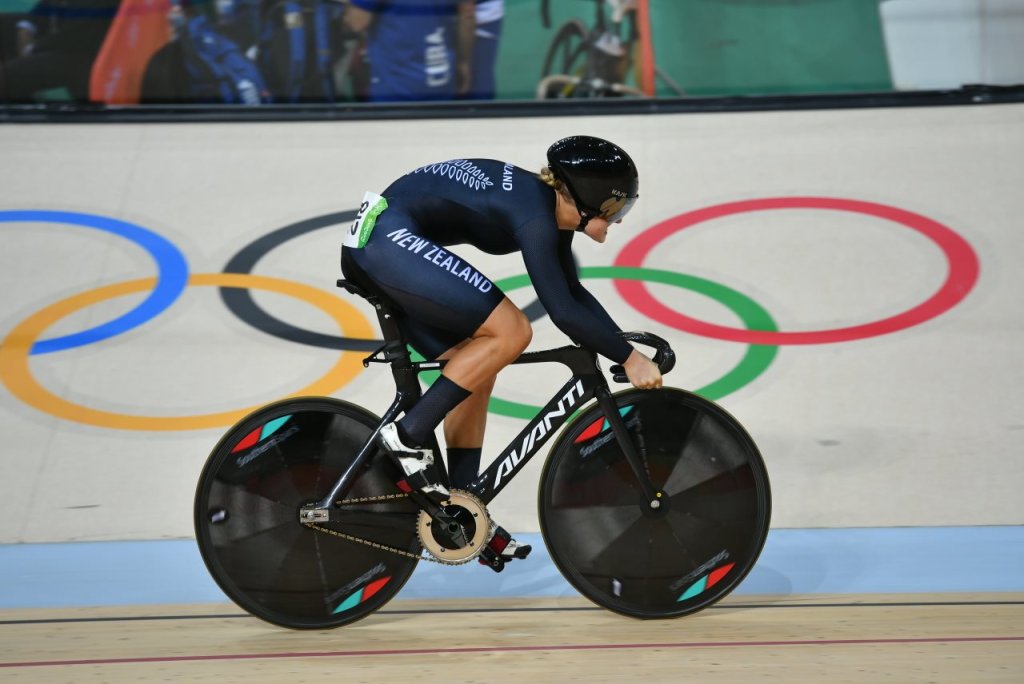 Ciclista durante competição olímpica; ela está em cima da bicicleta, em alta velocidade. O símbolo das Olimpíadas aparece ao fundo