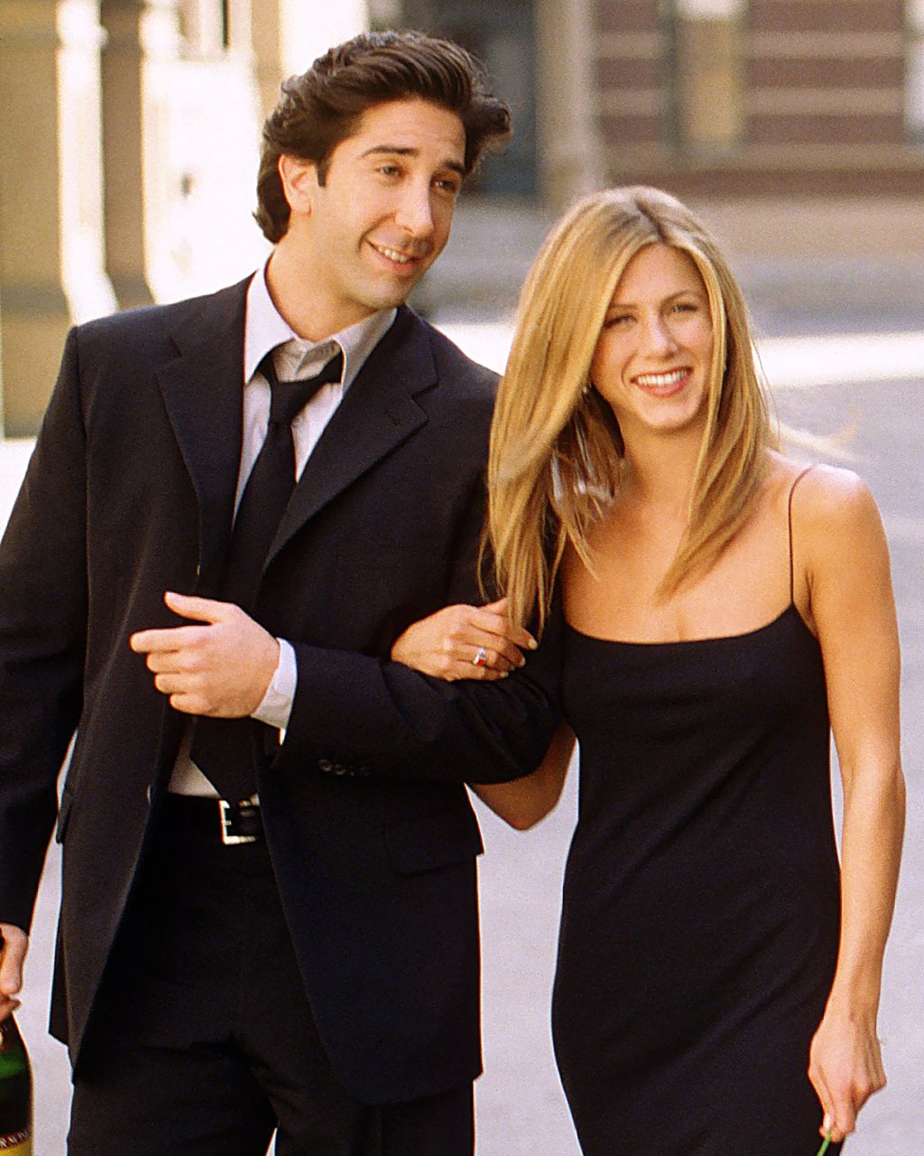 David Schwimmer e Jennifer Aniston como Ross e Rachel em Friends; eles estão usando roupas pretas e Jen está com o braço entrelaçado com o de David; os dois estão sorrindo para enquanto andam