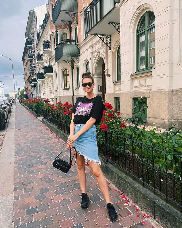 Jovem posando com o corpo inclinado, ela segura uma bolsa e usa camisa estampada com saia jeans e tênis.