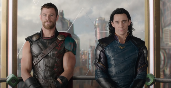 Thor e Loki em cena de Thor: Ragnarok; eles estão parados olhando para frente em um elevador de parede de vidro com as mãos na frente do corpo; Thor sorri levemente enquanto Loki tem uma expressão séria