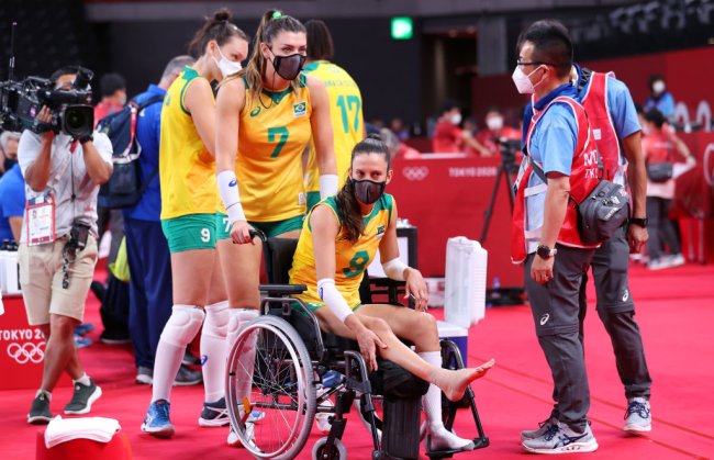 Rosamaria empurrando a cadeira de rodas da Macris, após a jogadora sofrer uma lesão no pé em partida contra o Japão em Tóquio. Macris está sentada, segurando a perna lesionada para cima.