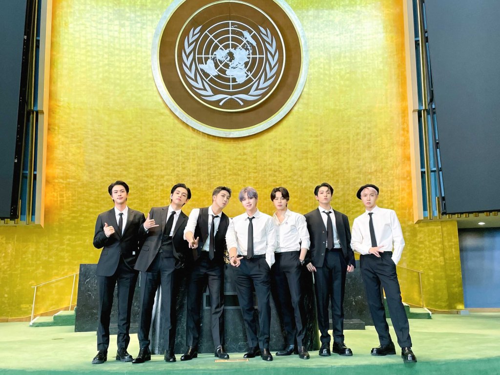 Os sete integrantes do BTS posando na frente do símbolo da ONU em uma parede dourada; eles usam terno e gravata preta com camisa branca