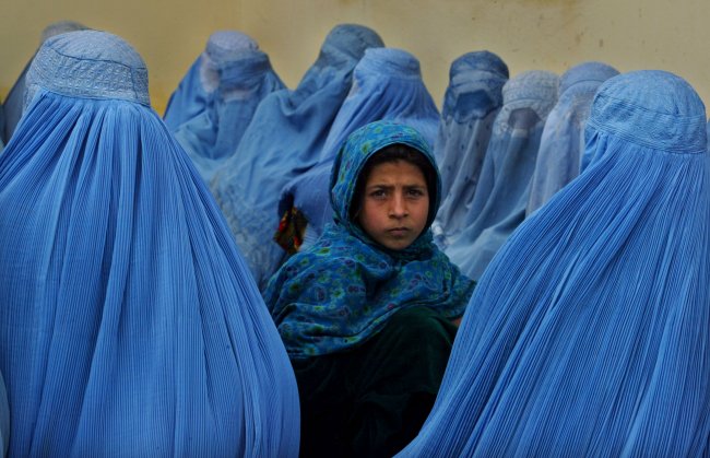 Mulheres afegãs vestem véu que cobre o rosto inteiro. Menina ao centro usa véu que cobre apenas o cabelo e olha para a câmera.