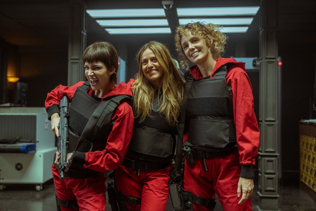 Úrsula Corberó, Itziar Ituño e Esther Acebo caracterizadas como Tokio, Lisboa e Estocolmo nos bastidores da parte 5 de La Casa de Papel; elas usam macacões vermelhos com colete à prova de balas preto e posam para câmera; Úrsula está segurando uma arma e fazendo careta enquanto Itziar e Esther sorriem para foto